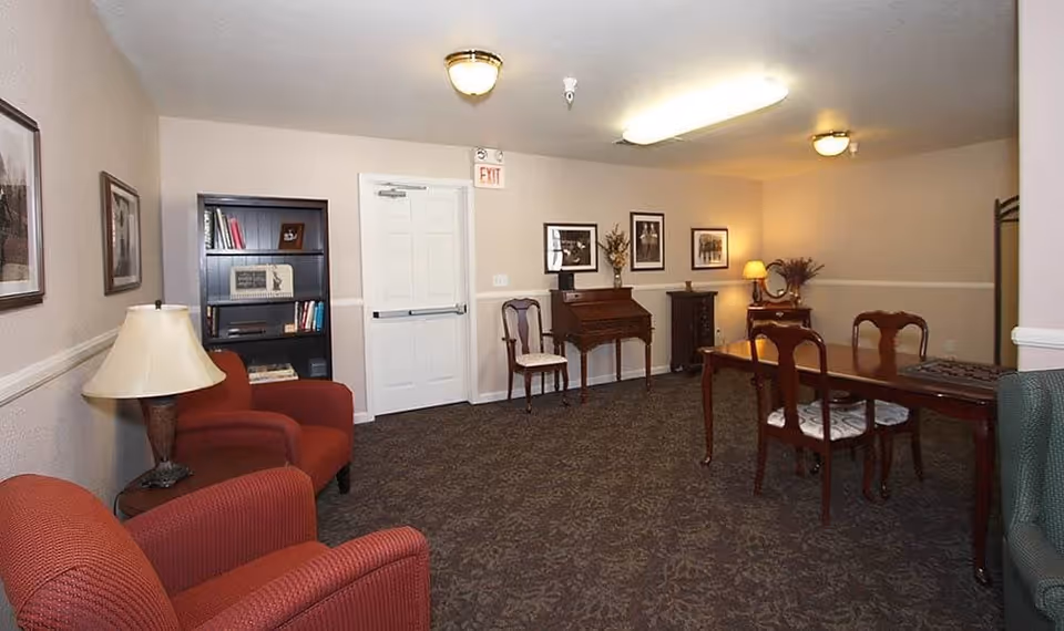 A cozy interior room with beige walls and patterned carpet. The room features two red armchairs with a lamp on a small table between them, a black bookshelf with books and decorations, a wooden desk with a chair, framed pictures on the walls, and a wooden dining table with chairs. The room is softly lit by ceiling lights and a table lamp.