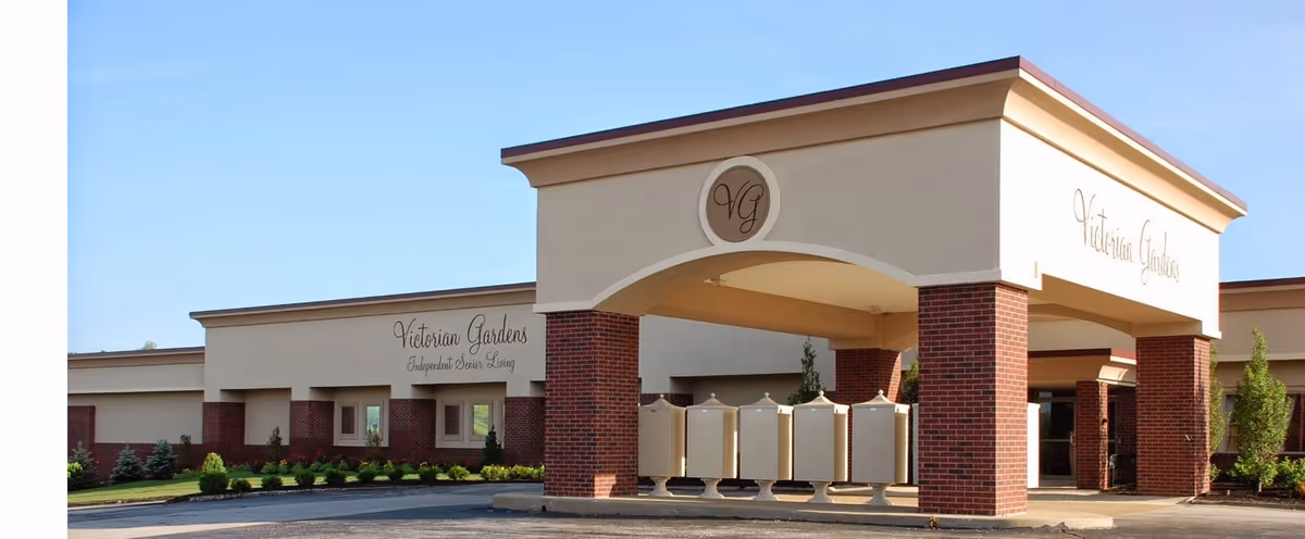 Exterior view of Victorian Gardens senior living facility showing the entrance with a covered drop-off area supported by brick columns, beige walls, and the facility name displayed on the building.