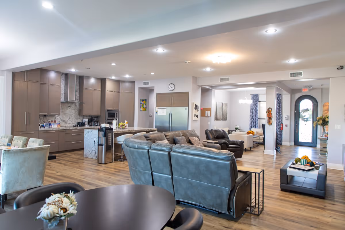 A spacious and modern open-concept living area in an assisted living facility featuring a kitchen with brown cabinets, a marble backsplash, and stainless steel appliances. The living room area has leather sofas and armchairs arranged around a coffee table. There is a dining area with upholstered chairs and a round table in the foreground. The room has wooden flooring, recessed lighting, and a glass front door with a wreath.