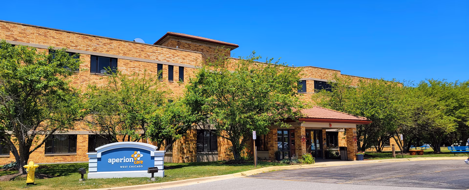 Exterior view of Aperion Care West Chicago facility showing a two-story brick building with multiple windows, a covered entrance with a red-tiled roof, surrounded by green trees and a clear blue sky. A sign with the facility name is visible on the lawn near the entrance.