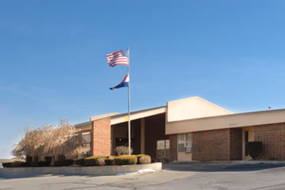 Exterior view of a single-story brick building with a covered entrance, two flagpoles displaying the American flag and another flag, and some landscaping including bushes and a tree. The sky is clear and blue.