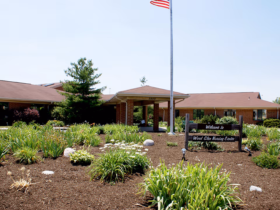 Exterior view of Wood Glen Nursing Center showing a single-story brick building with a covered entrance, surrounded by landscaped garden beds with green plants and flowers. An American flag is flying on a flagpole near the entrance, and a wooden sign reads 'Welcome to Wood Glen Nursing Center'.