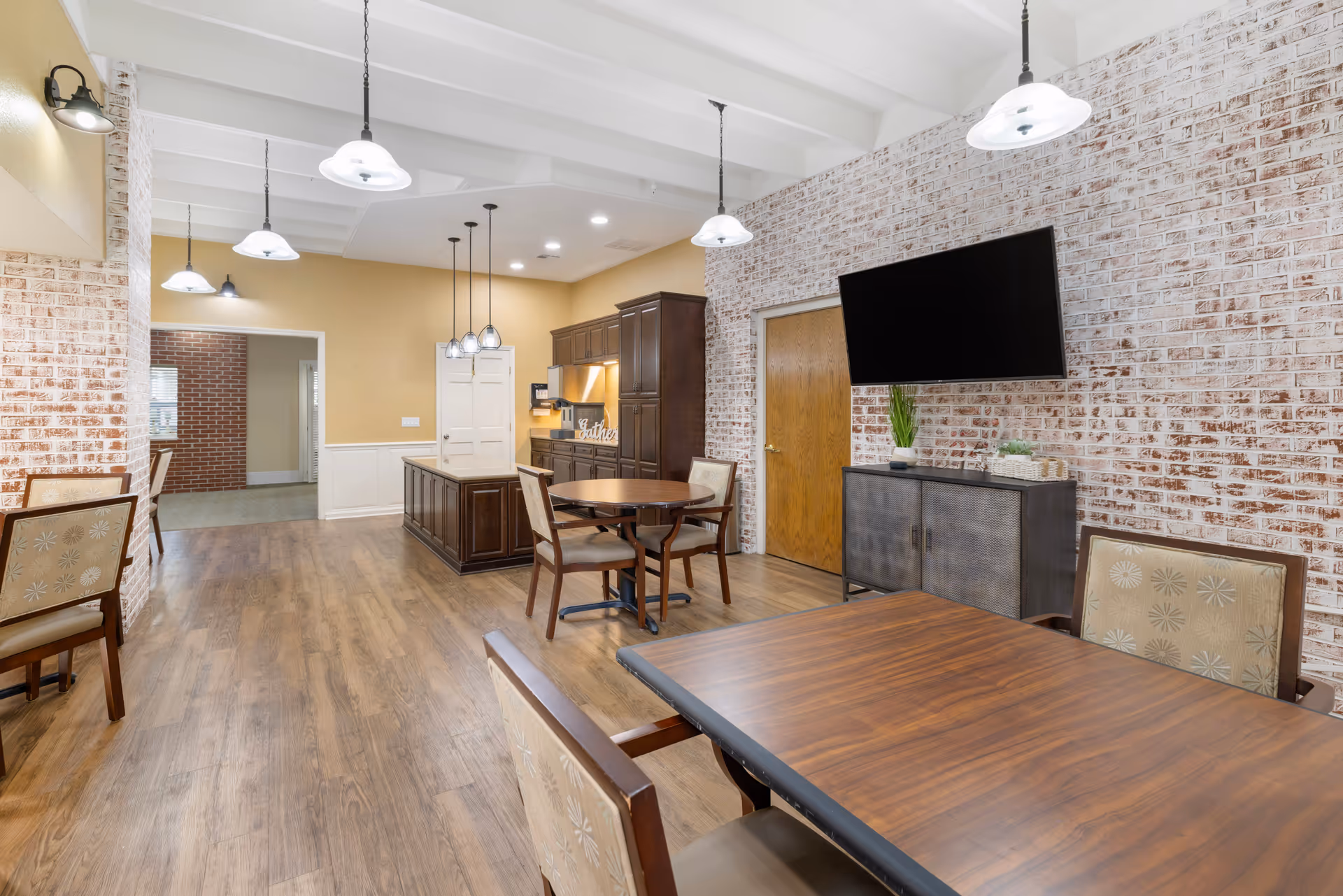 Interior view of a senior living facility dining area with wooden tables and cushioned chairs. The room features a whitewashed brick wall with a mounted flat-screen TV above a dark cabinet. Pendant lights hang from a white beamed ceiling, and a kitchen area with dark wood cabinets and an island is visible in the background.