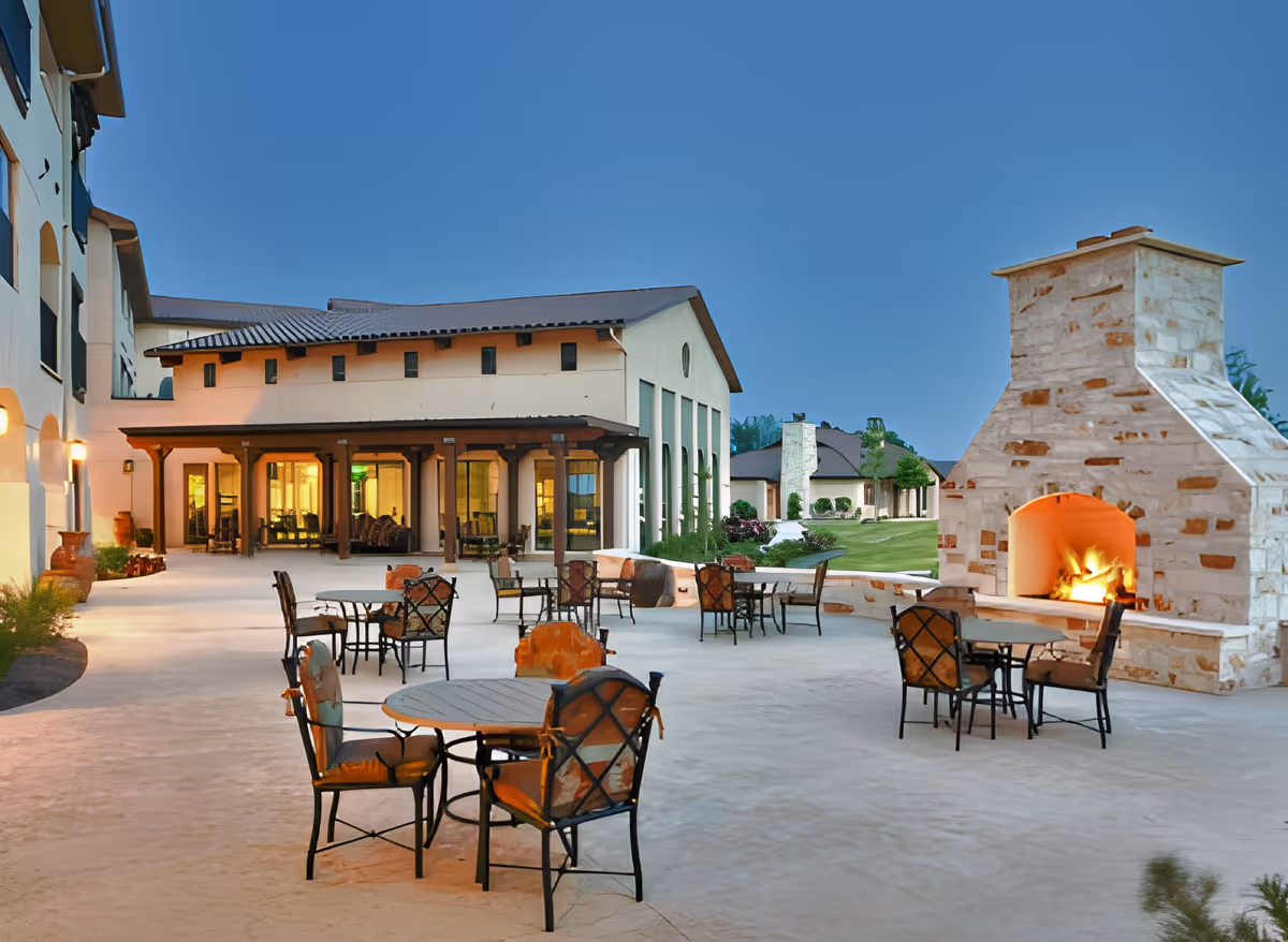 Outdoor patio area at Buckner Villas featuring multiple round tables with cushioned chairs, a large stone fireplace with a fire burning, and a building with large windows and a covered porch in the background under a clear evening sky.