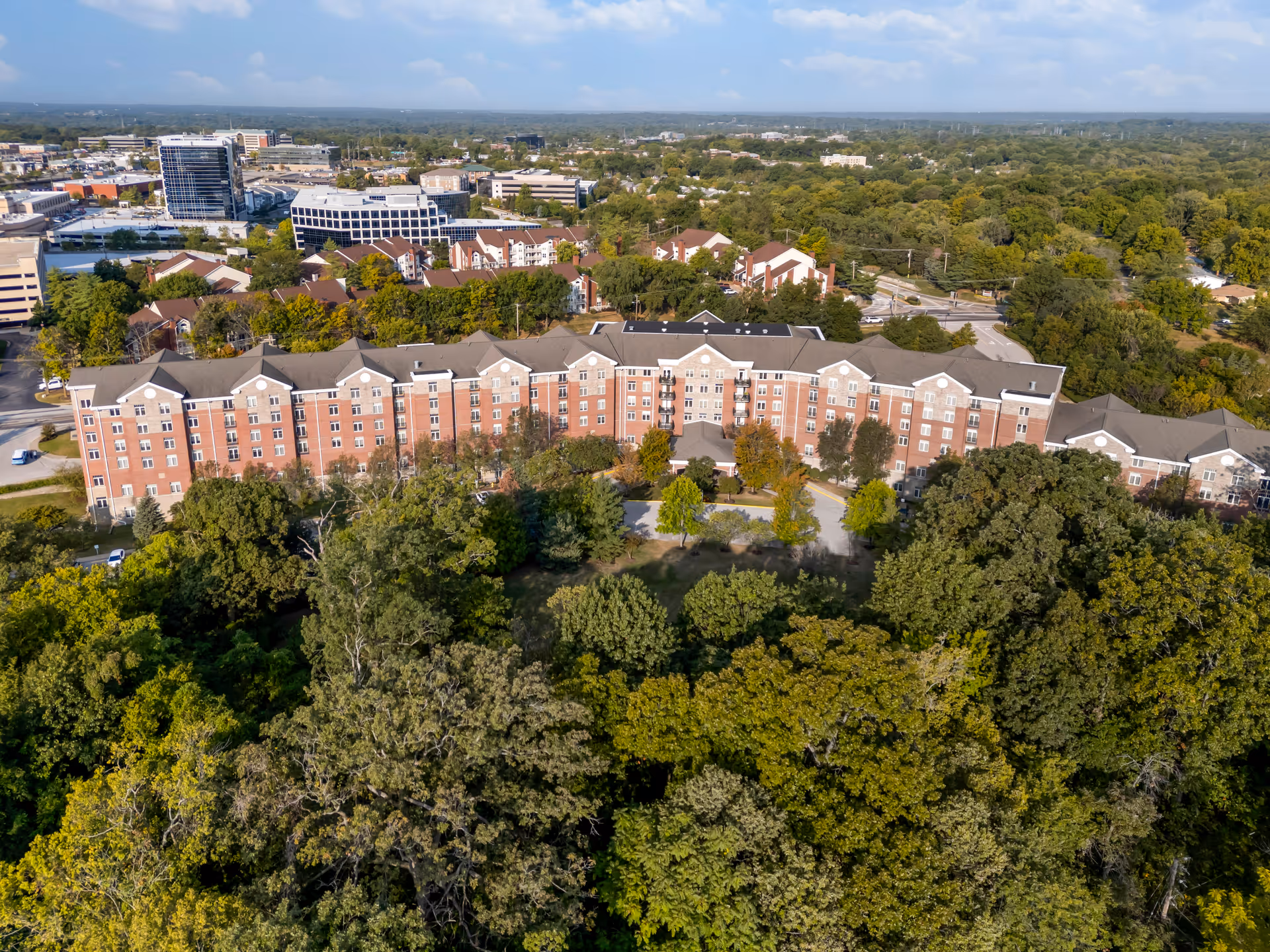 Aerial view of a large multi-story senior living facility building surrounded by dense green trees and landscaping, with a cityscape visible in the background under a partly cloudy sky.