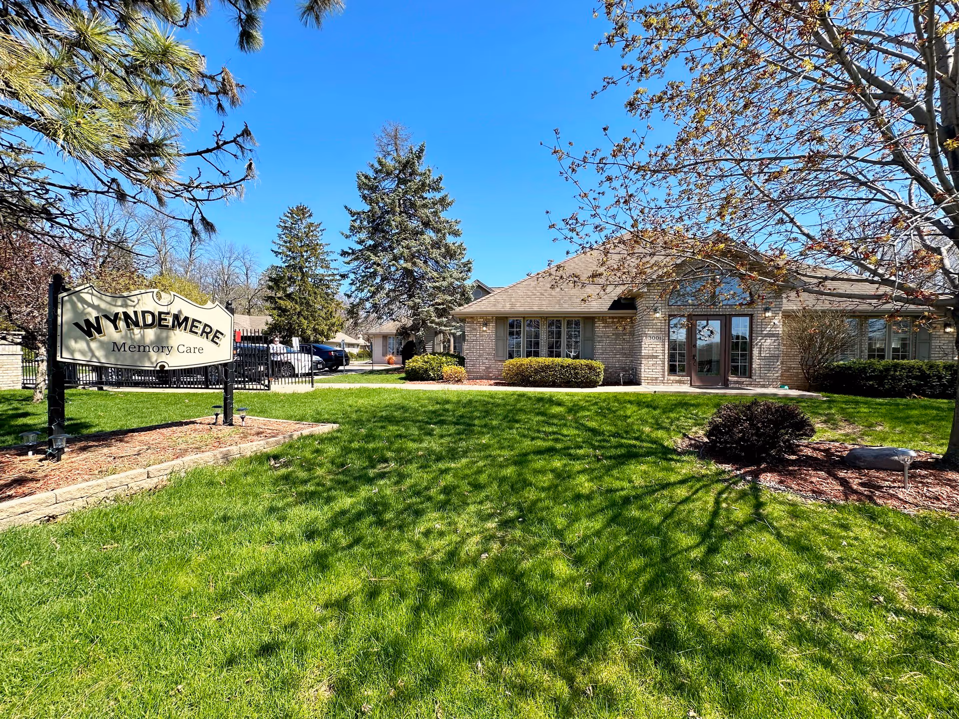 Exterior view of Wyndemere Memory Care facility with a well-maintained green lawn, trees, and a clear blue sky. A sign with the facility name is visible on the left side, and the building with large windows and a main entrance is on the right.
