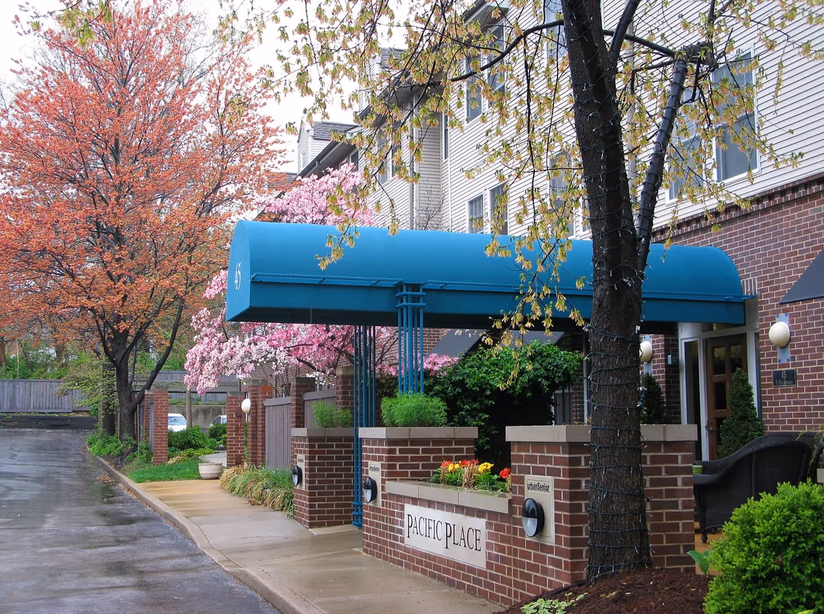 Entrance to Pacific Place Senior Living facility with a blue canopy over the walkway, brick pillars, and a sign reading 'PACIFIC PLACE'. Surrounding the entrance are blooming trees with pink and orange flowers, green shrubs, and a wet pavement indicating recent rain.