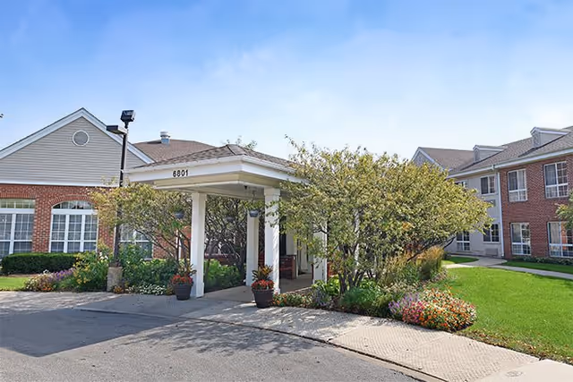 Exterior view of a senior living facility entrance with a covered drop-off area supported by white columns, surrounded by green shrubs, flowering plants, and trees. The building features red brick and beige siding with multiple windows and a well-maintained lawn.