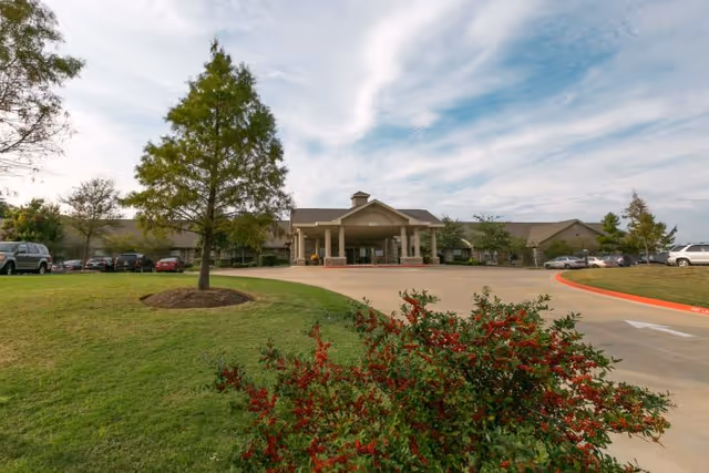 Front exterior view of Legend Oaks Healthcare and Rehabilitation - North Austin building with a driveway, landscaped lawn, trees, and parked cars under a partly cloudy sky.