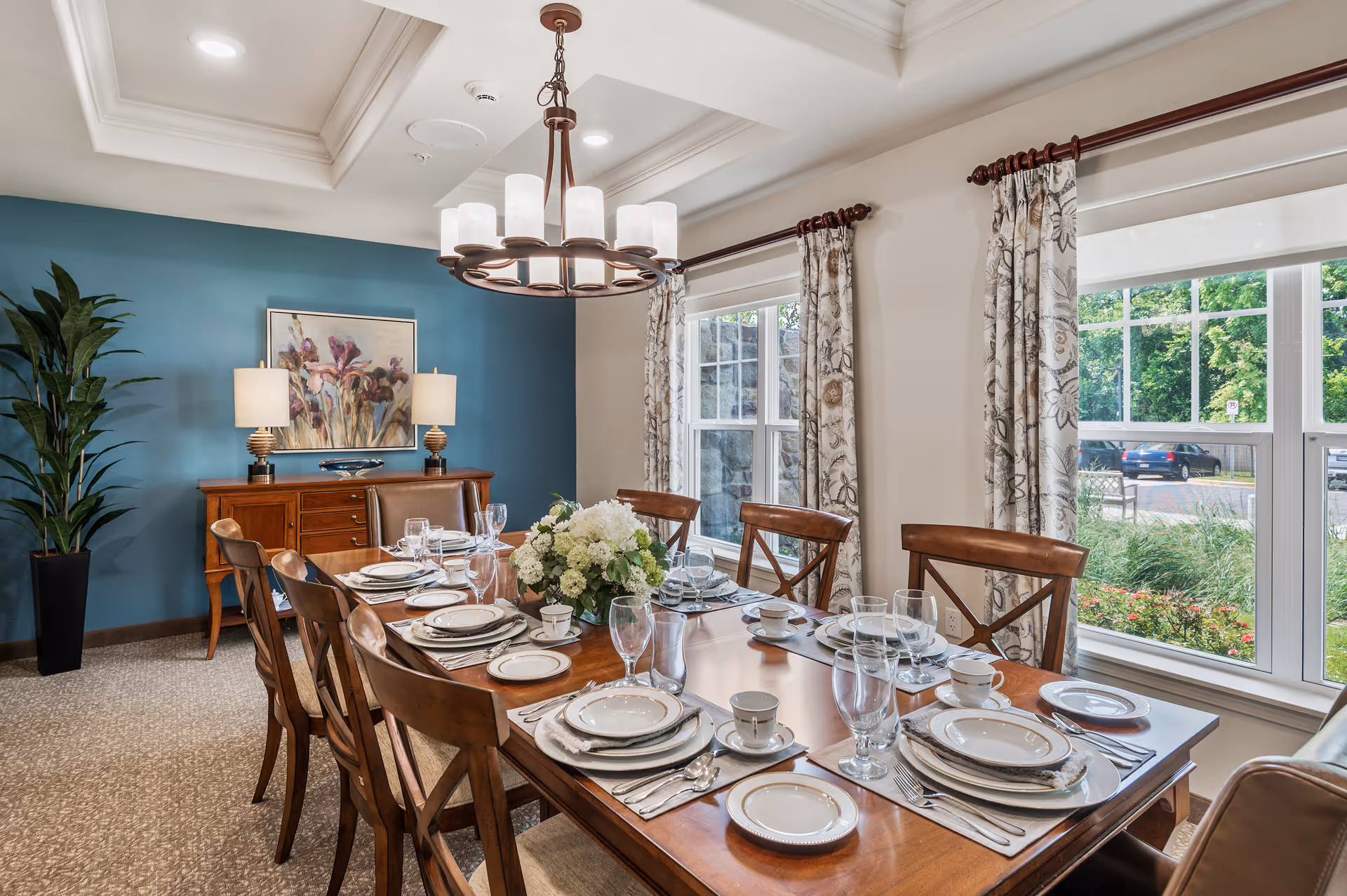 A well-lit dining room with a wooden dining table set for eight people, featuring white plates, cups, and glassware. The room has large windows with floral curtains allowing natural light to brighten the space. A blue accent wall with a painting and two table lamps on a wooden sideboard is visible, along with a tall green plant in a black pot in the corner.