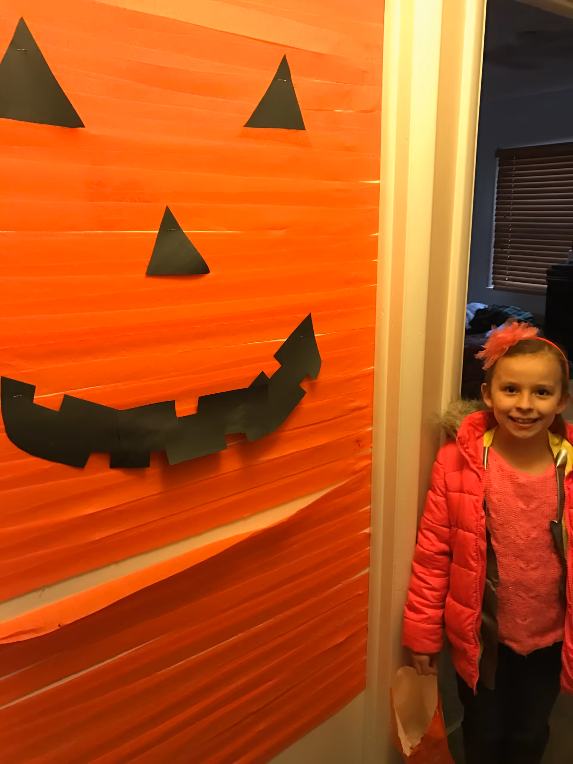 A young girl wearing a pink jacket and a pink headband stands smiling in a doorway next to a wall decorated with an orange paper pumpkin face made from black paper cutouts for eyes, nose, and mouth.