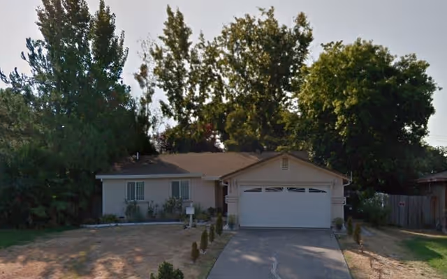 Single-story residential house with a two-car garage, surrounded by tall trees and a driveway leading up to the garage. The lawn appears dry with some small shrubs near the house.