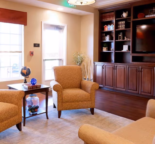 A cozy living room area with beige upholstered armchairs and a matching sofa arranged around a wooden side table. The side table holds a globe, a blue decorative orb, and some books. Behind the seating area is a large built-in wooden cabinet with shelves displaying decorative items and a flat-screen TV. A window with a red valance and a door marked 'EXIT' are visible in the background, along with a vase of flowers and a light fixture on the ceiling.