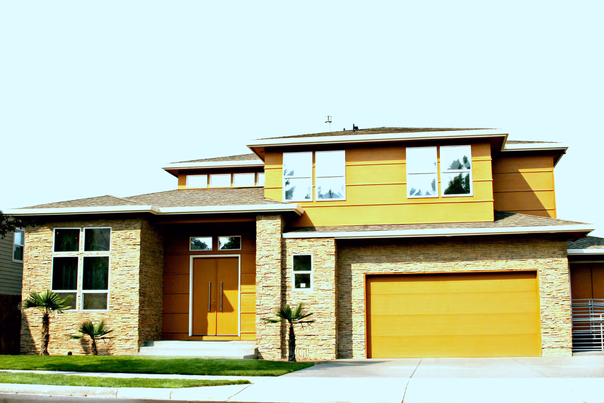 Front exterior view of a modern two-story house with a combination of stone and yellow siding, large windows, a double front door, a two-car garage, and small palm trees in the front yard.