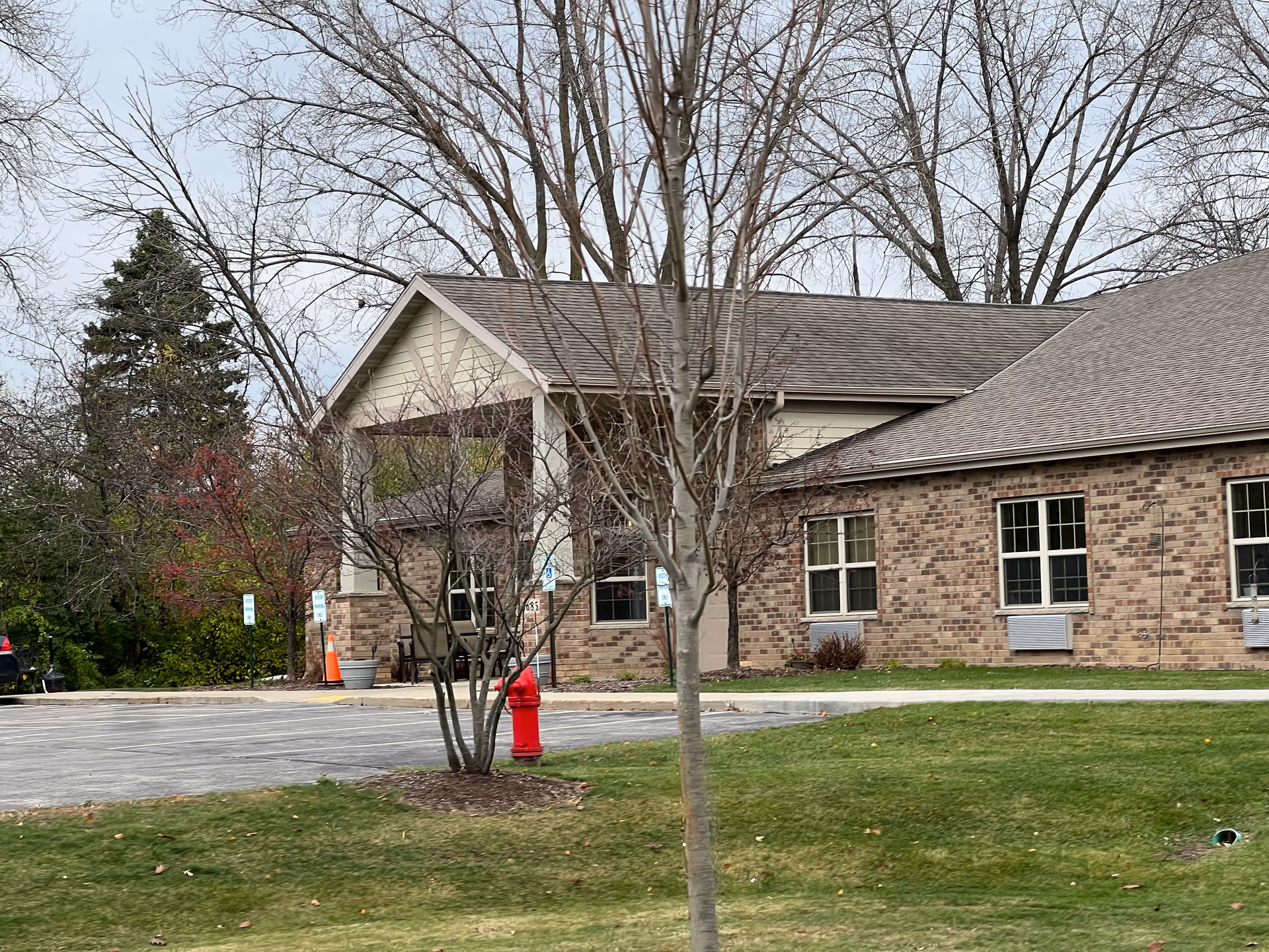 Exterior view of a single-story brick building with a gabled roof, surrounded by leafless trees and a grassy area. There is a parking lot with handicap parking signs and a red fire hydrant near the entrance.