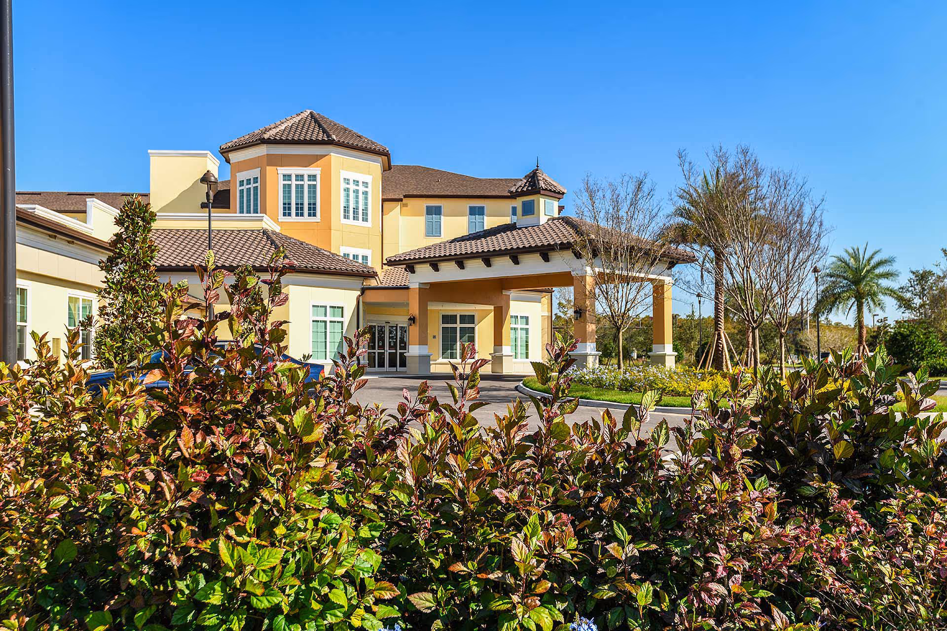 Exterior view of a senior living facility building with a covered entrance, surrounded by landscaped greenery and trees under a clear blue sky.