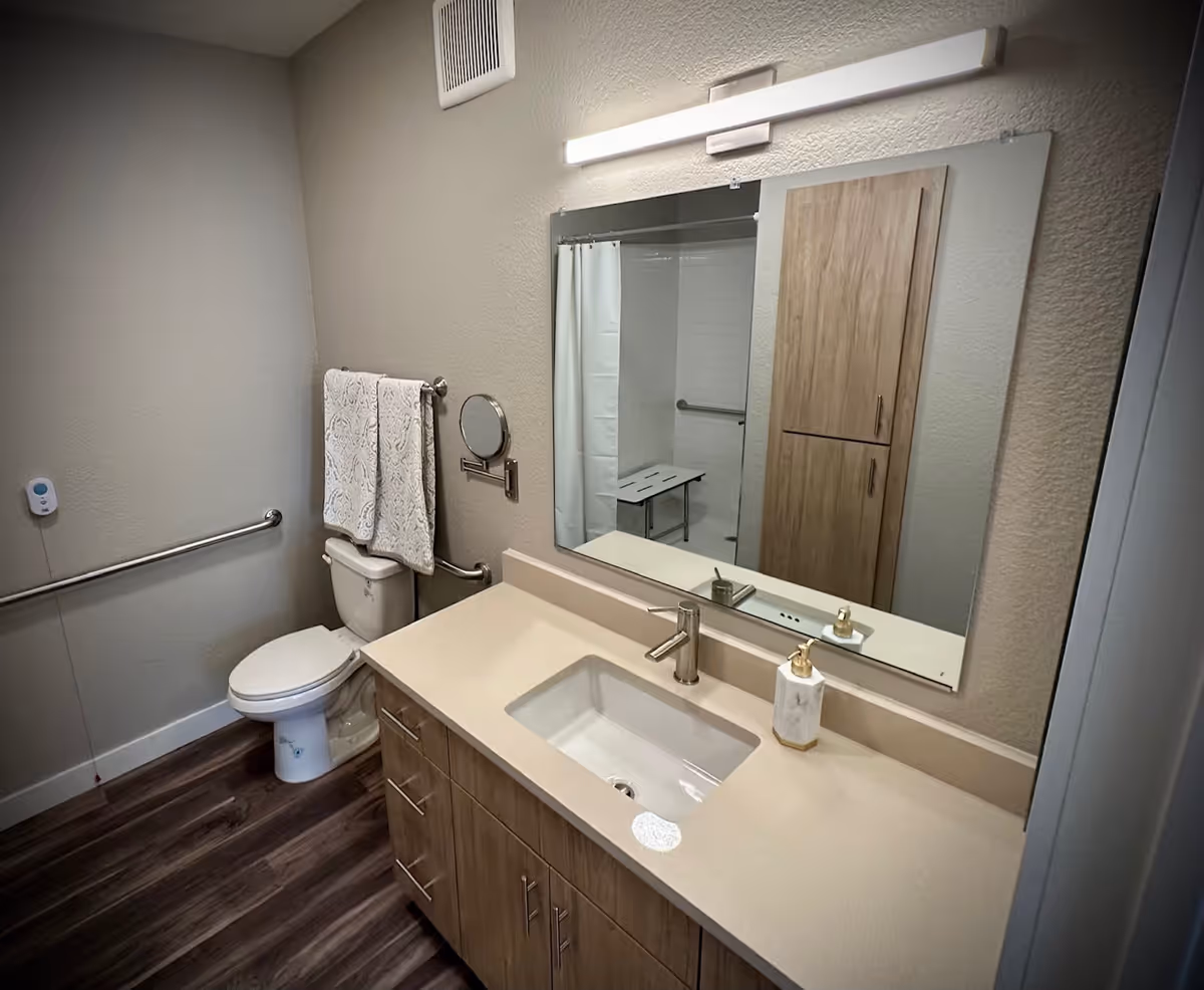 A modern bathroom featuring a sink with a mirror, a toilet, and wooden cabinetry. The space is well-lit with a towel hanging on a rack and a shower area visible in the background.