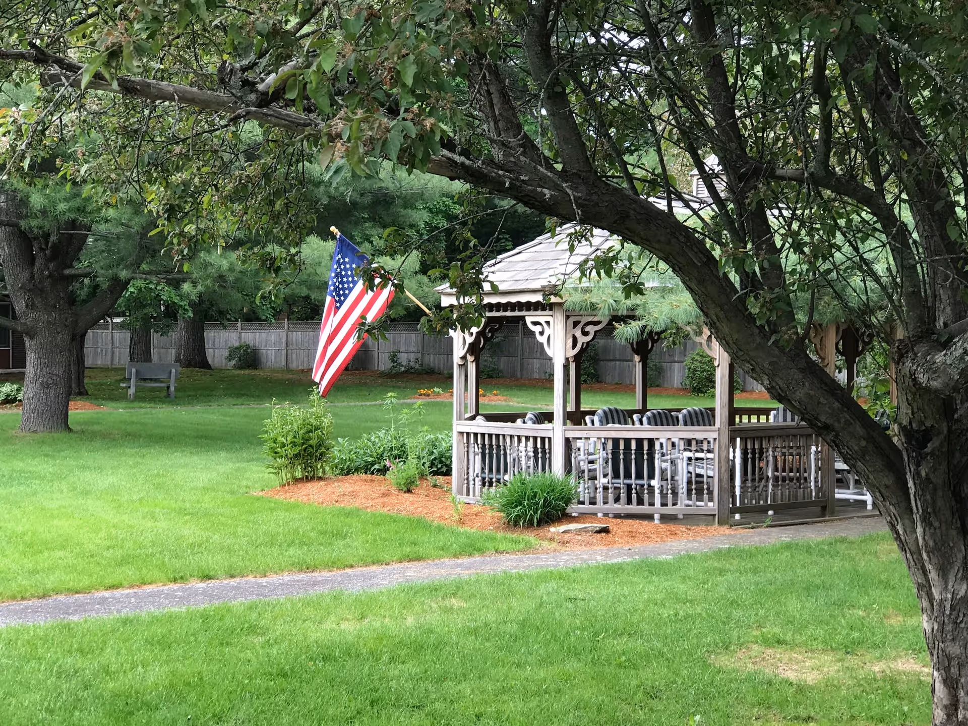 A peaceful outdoor garden area with a wooden gazebo surrounded by green grass and trees. An American flag is displayed near the gazebo, and there is a wooden bench in the background near a wooden fence.