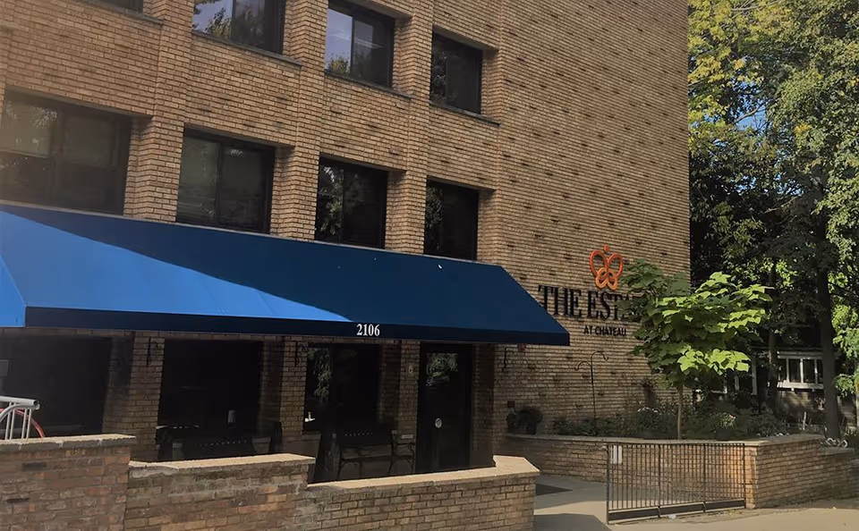 Exterior view of a brick building with a blue awning over the entrance. The building has several windows and a sign on the wall that reads 'The Estates at Chateau' with an orange butterfly logo. There are trees and greenery around the entrance area.