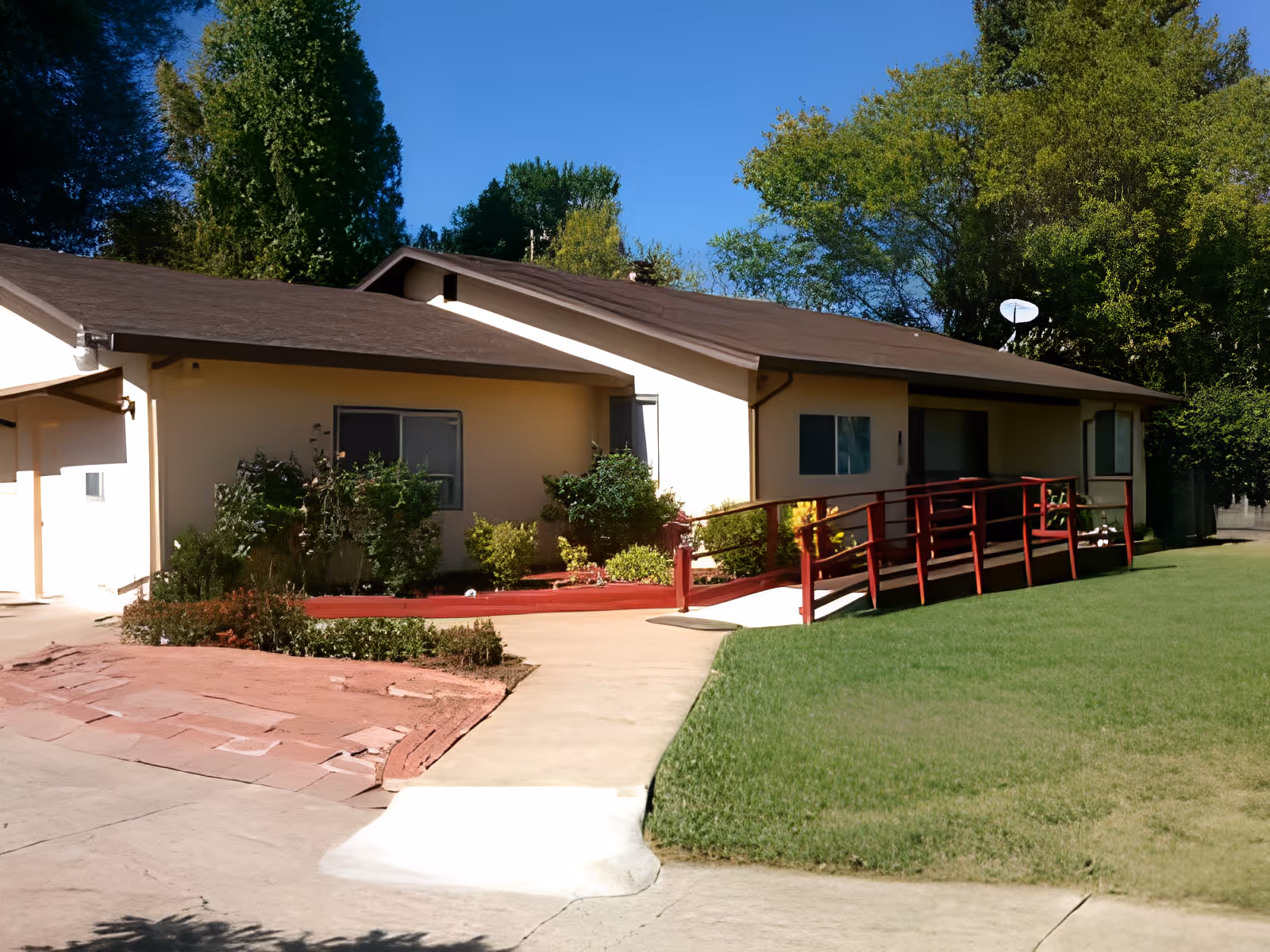 Single-story building with beige walls and a brown roof, surrounded by green trees and bushes. There is a concrete pathway leading to a red wooden ramp with railings, providing wheelchair access to the entrance. The sky is clear and blue.