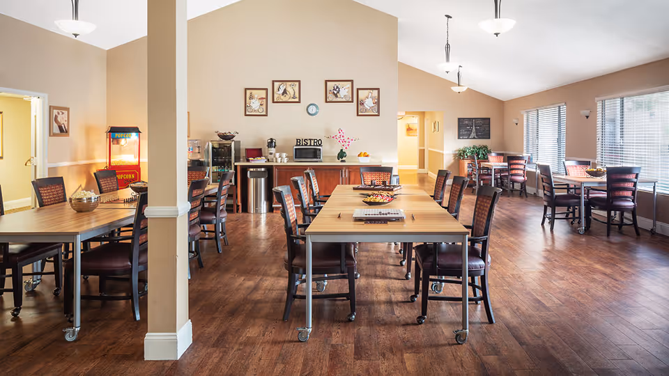 A bright dining area with multiple tables and chairs arranged neatly on a wooden floor. The room features large windows with blinds allowing natural light to fill the space. On the left side, there is a popcorn machine and a counter with a microwave, coffee maker, and decorative items. The walls are adorned with framed pictures and a clock, and the ceiling has hanging light fixtures.