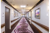 A long, well-lit hallway in a skilled nursing and rehabilitation center with patterned carpet in shades of purple and brown. The hallway has handrails on both sides, several closed doors, and framed artwork hanging on the walls.