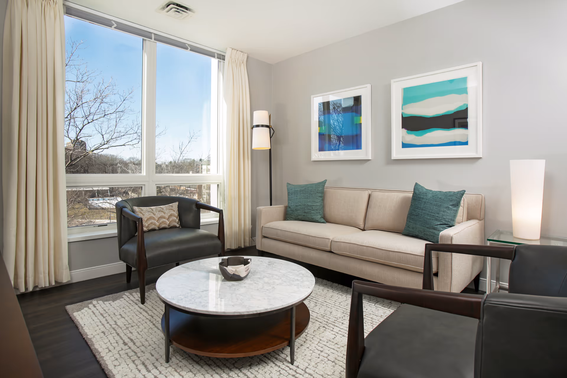 A modern living room with a beige sofa adorned with two teal pillows, two black armchairs with wooden arms, a round marble coffee table on a textured rug, a tall floor lamp, and two abstract framed artworks on a light gray wall. Large windows with cream curtains allow natural light to fill the room, showing a view of trees outside.