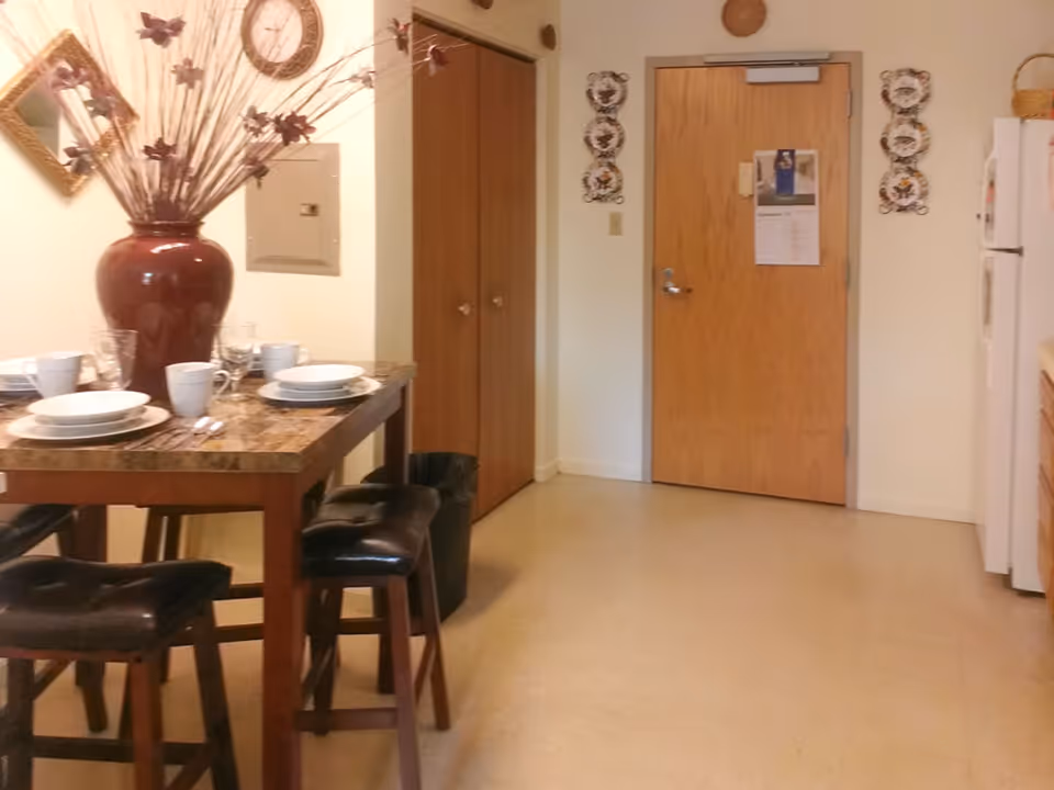 Interior view of a dining area in an apartment with a wooden table set for four people with white plates, cups, and glasses. A large decorative vase with dried flowers is on the table. There are four dark cushioned stools around the table. In the background, there is a wooden door with a calendar hanging on it, two wooden closet doors, and a white refrigerator to the right.