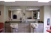Interior view of a salon area within a nursing and rehabilitation center featuring two salon sinks with chairs, large mirrors above the sinks, cabinets and drawers underneath, and a fire extinguisher mounted on the wall to the right.
