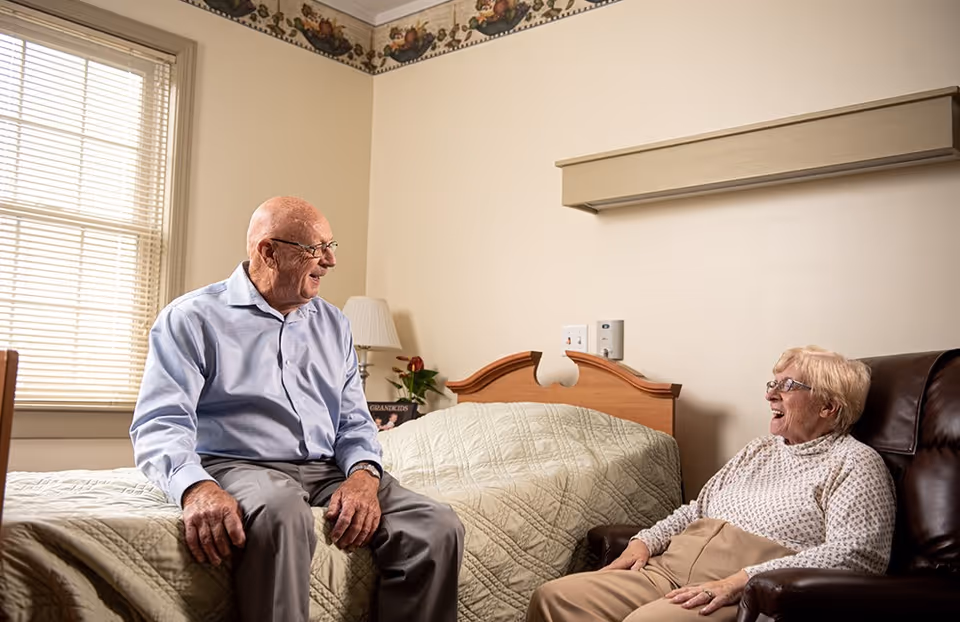 An elderly man and woman smiling and conversing in a cozy room. The man is sitting on the edge of a bed with a beige quilt, and the woman is seated in a brown leather chair. The room has beige walls, a window with blinds, a lamp, and a small plant on a nightstand.