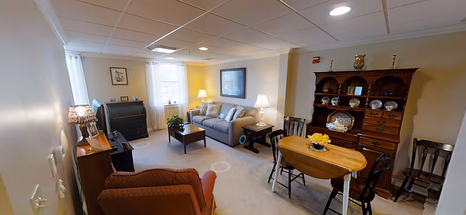 A cozy living room with a beige sofa, two side tables with lamps, a wooden coffee table, an armchair, a small wooden dining table with chairs, and a wooden hutch displaying decorative plates and items. The room has two windows with sheer white curtains and light-colored walls.