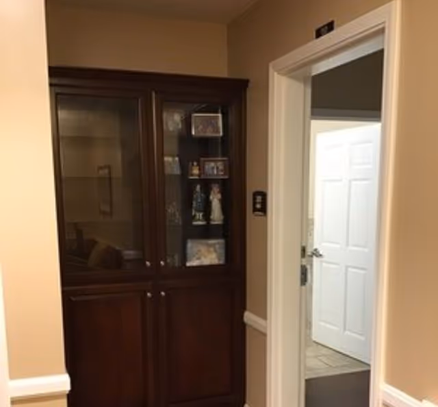 Interior view of a hallway corner in a senior living facility with beige walls and white trim. A dark wooden cabinet with glass doors displays decorative items and photos. An open doorway reveals a white door and tiled floor in the adjacent room.