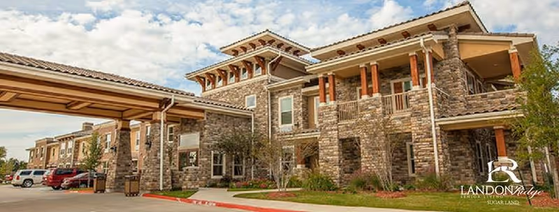 Exterior view of a senior living facility building with stone and wood architectural elements, a covered entrance, and several parked cars under a partly cloudy sky.