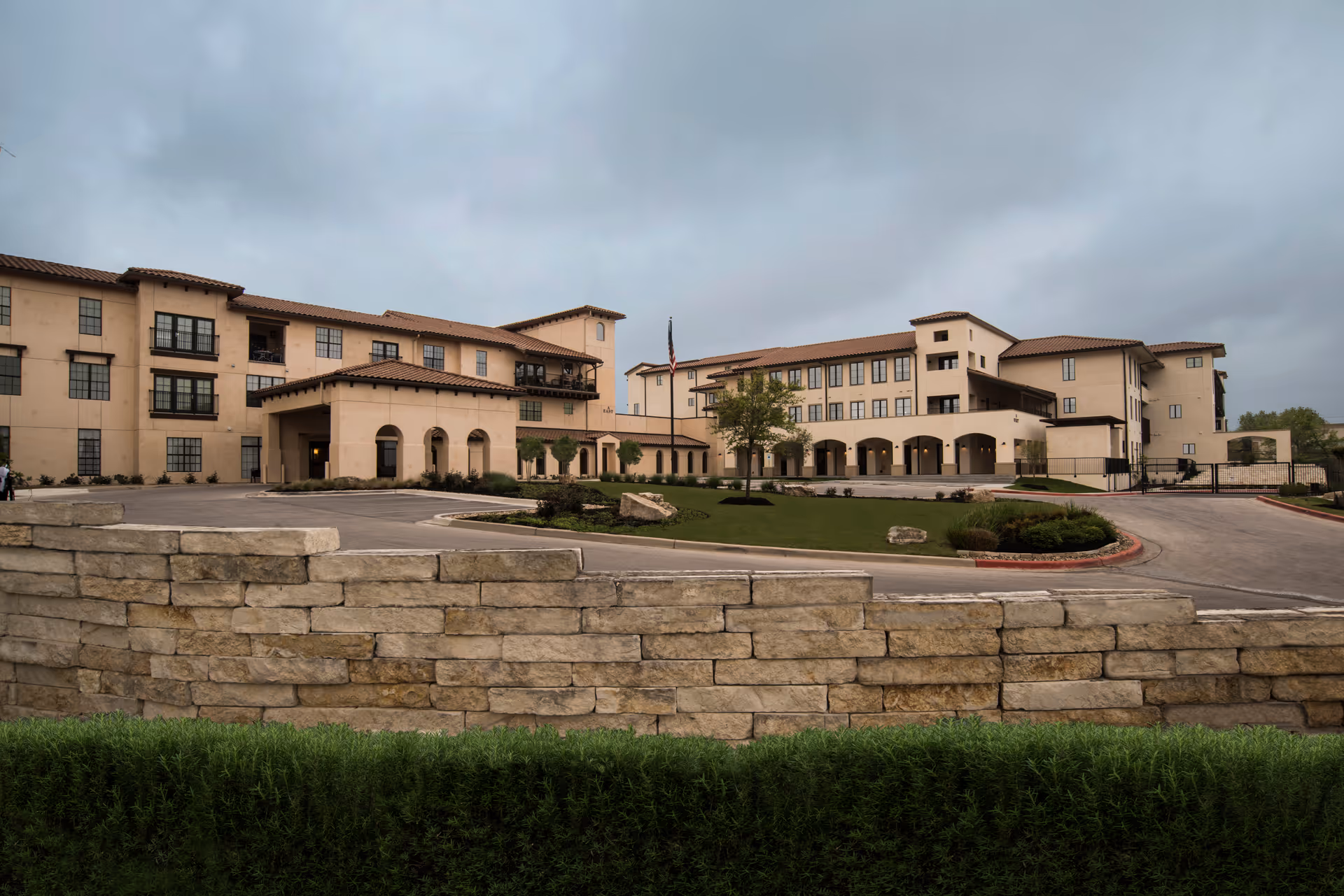 Exterior view of Buckner Villas, a multi-story senior living facility with beige walls and a red-tiled roof. The building is surrounded by a stone wall and landscaped greenery, with a driveway leading to the entrance under a cloudy sky.