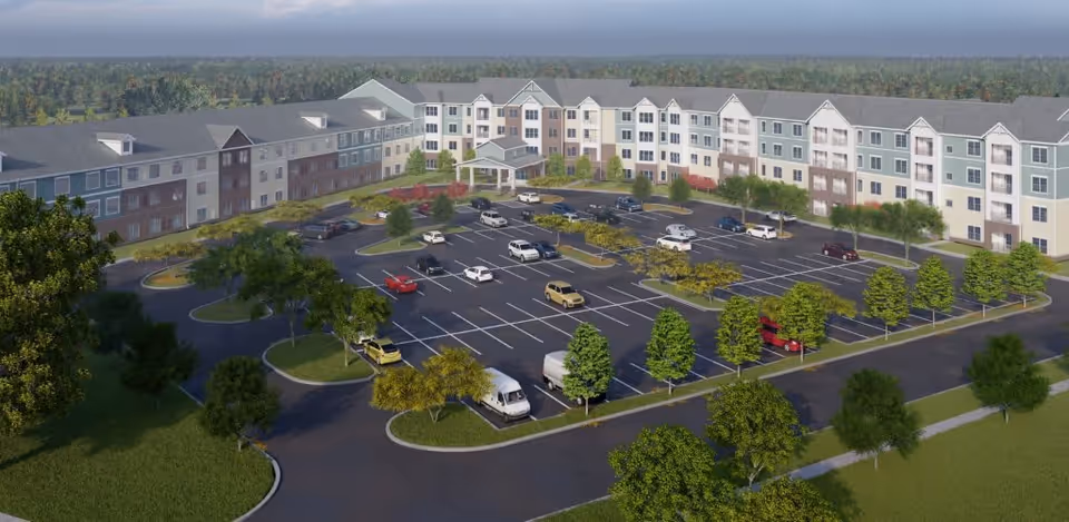 A large, multi-story senior living facility named Harmony at Avon with a spacious parking lot in front, surrounded by landscaped greenery and trees under a clear sky.