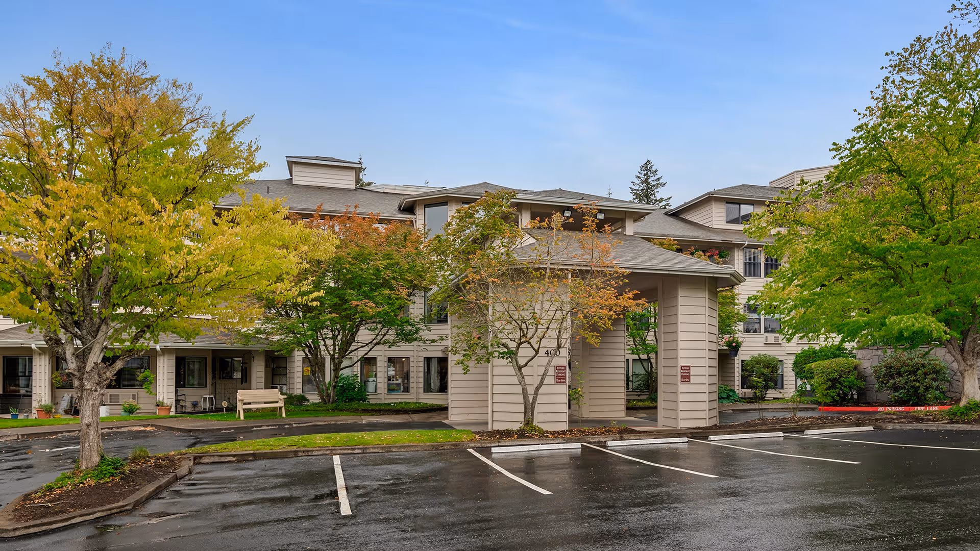 Exterior view of Holiday Hidden Lakes senior living facility showing a multi-story building with beige siding, surrounded by trees with green and autumn-colored leaves. There is a covered entrance with a driveway and empty parking spaces in the foreground on a wet pavement.