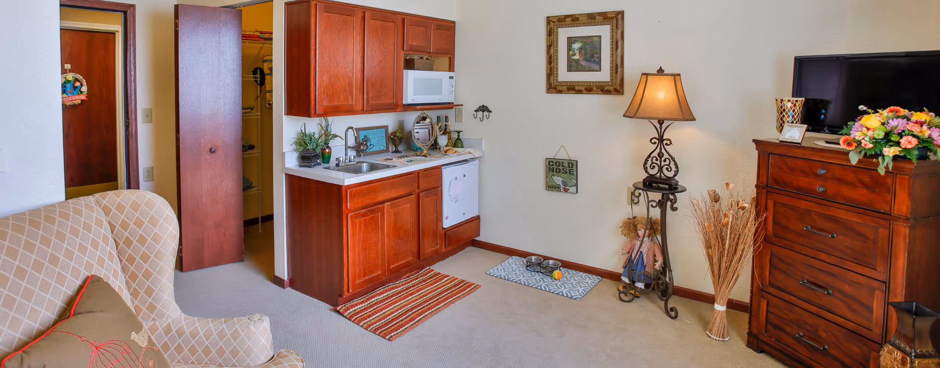 Interior view of a senior living facility room featuring a small kitchenette with wooden cabinets, a sink, microwave, and dishwasher. To the left is a beige armchair with a decorative pillow. On the right side, there is a wooden dresser with a TV on top, a floor lamp, and decorative items including a vase with dried plants. The room has light-colored walls and carpeted floor.