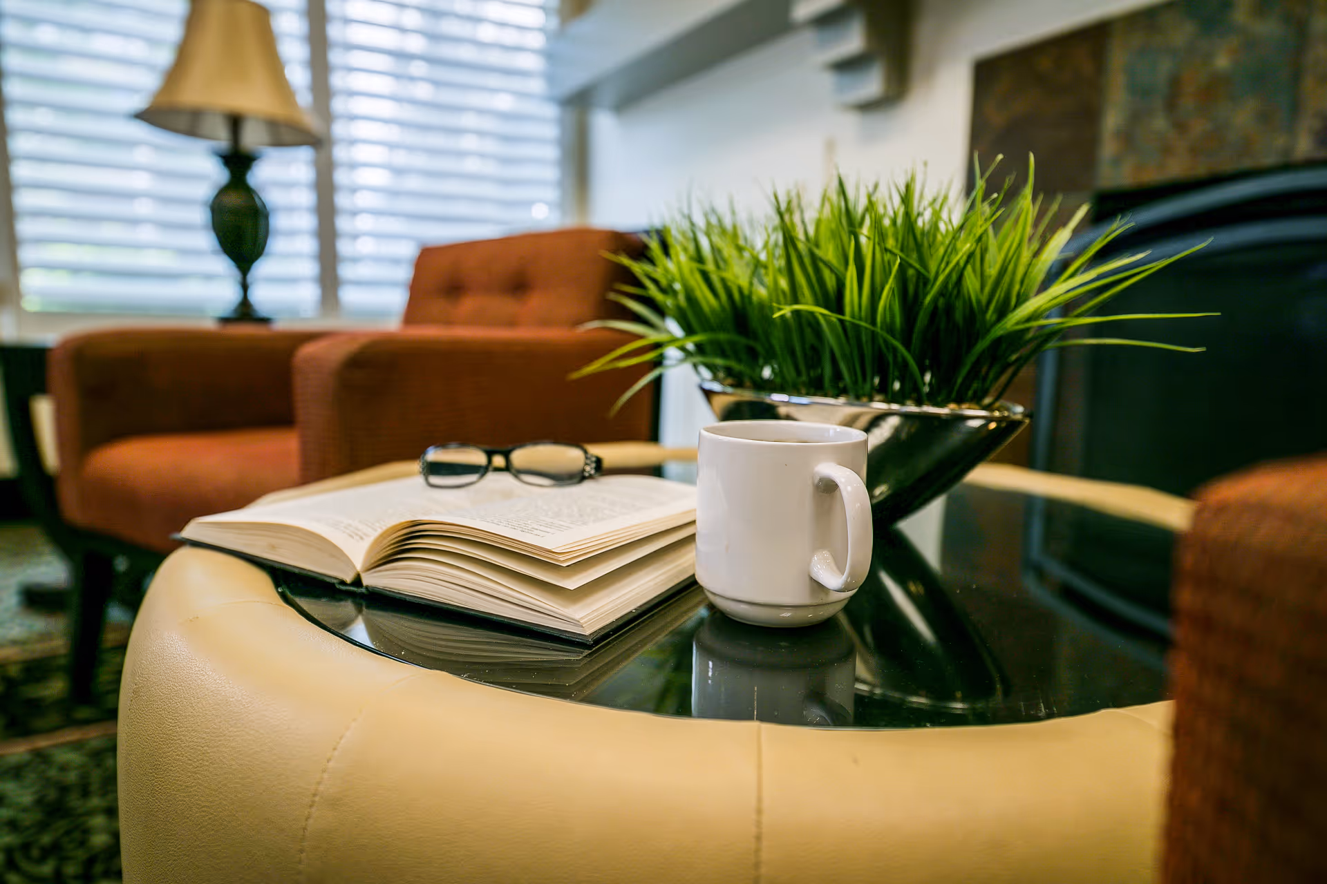 A cozy living room setting with a glass-top coffee table holding an open book, a pair of eyeglasses, a white coffee mug, and a decorative green plant. In the background, there are two brown upholstered armchairs and a lamp near a window with blinds.