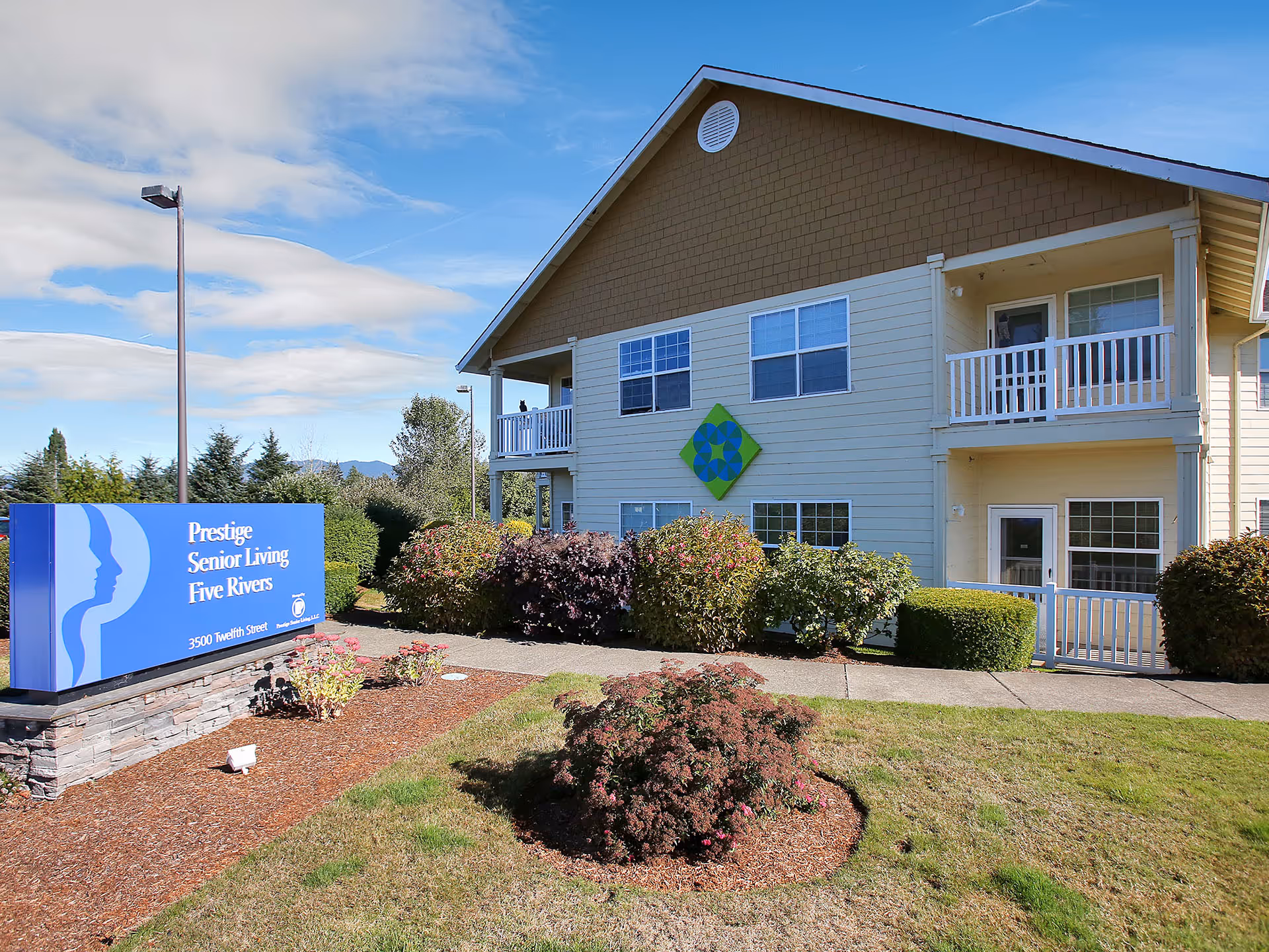Exterior view of Prestige Senior Living Five Rivers building with a blue sign displaying the facility name and address. The building is two stories with balconies and surrounded by bushes and a well-maintained lawn under a partly cloudy sky.