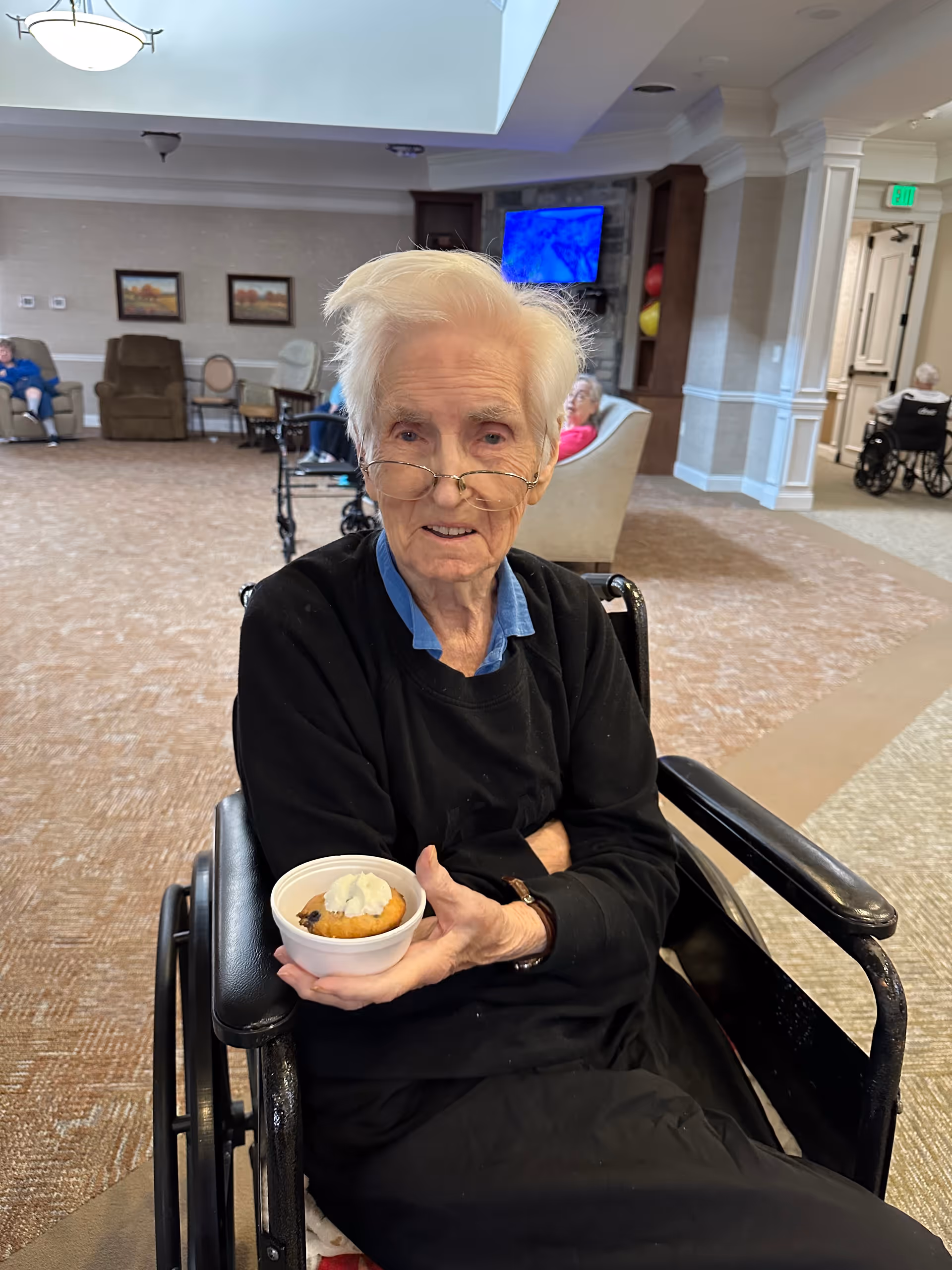 An elderly person with white hair and glasses sitting in a wheelchair inside a senior living facility common area, holding a small dessert cup with whipped cream. The room has carpeted floors, chairs, a TV mounted on the wall, and other elderly residents in the background.