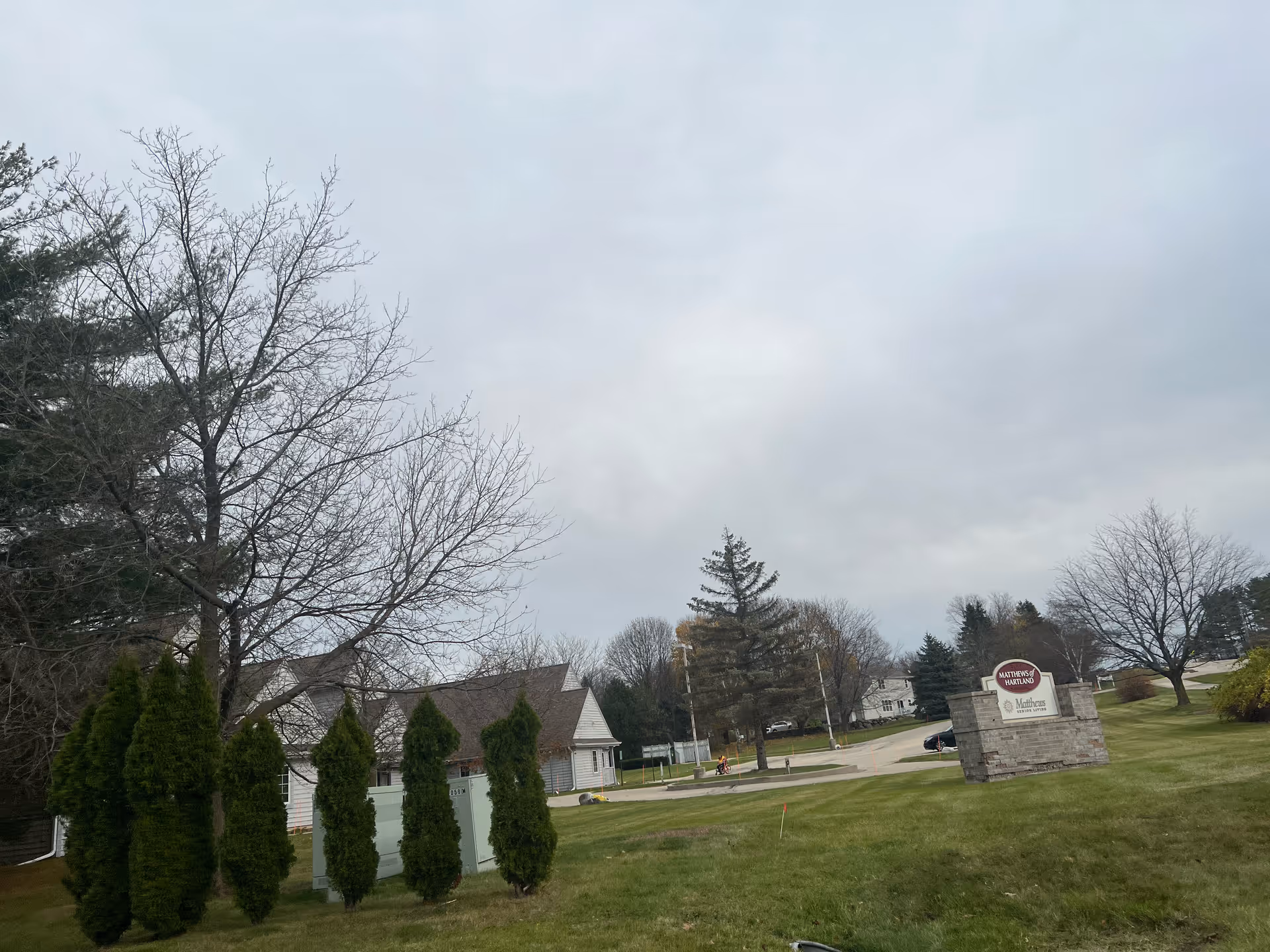 Outdoor view of Matthews Of Hartland facility with a stone sign displaying the facility name on a grassy lawn, surrounded by trees and a cloudy sky.