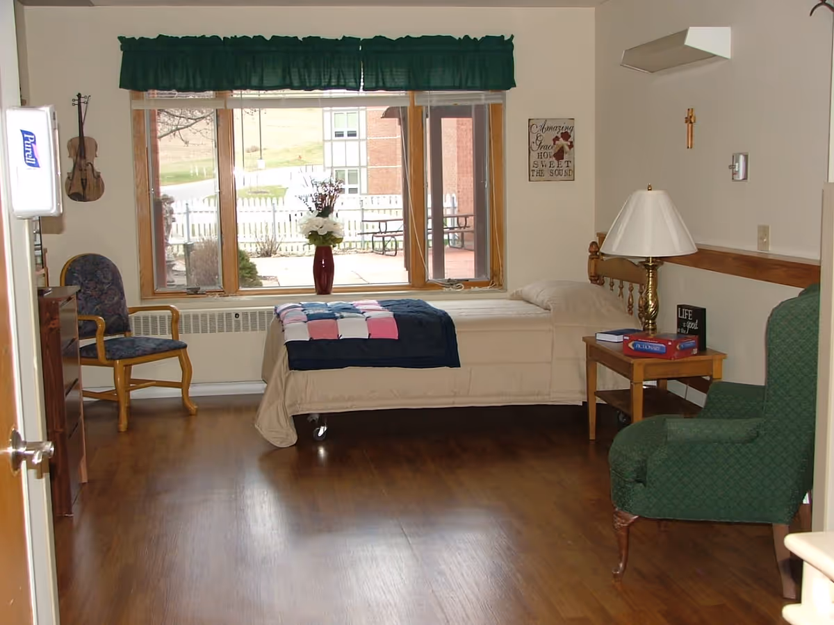 A cozy bedroom in a retirement community featuring a single bed with a beige comforter and a colorful patchwork blanket. There is a wooden nightstand with a lamp and board games, a green upholstered armchair, a wooden chair with a patterned cushion, and a large window with a green valance overlooking an outdoor area with a white picket fence and picnic tables. The room has wood flooring and light-colored walls with decorative wall hangings.