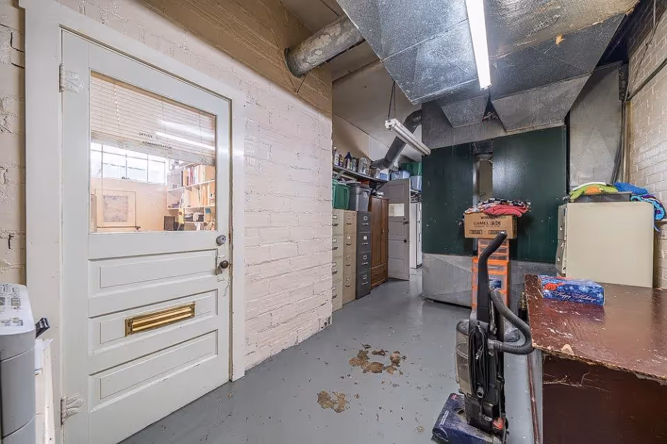A storage or utility room with a white door labeled Office Administration, beige brick walls, a gray concrete floor with some stains, metal ductwork on the ceiling, filing cabinets, shelves with various items, a vacuum cleaner, and a wooden desk with some folded clothes and a tissue box on it.