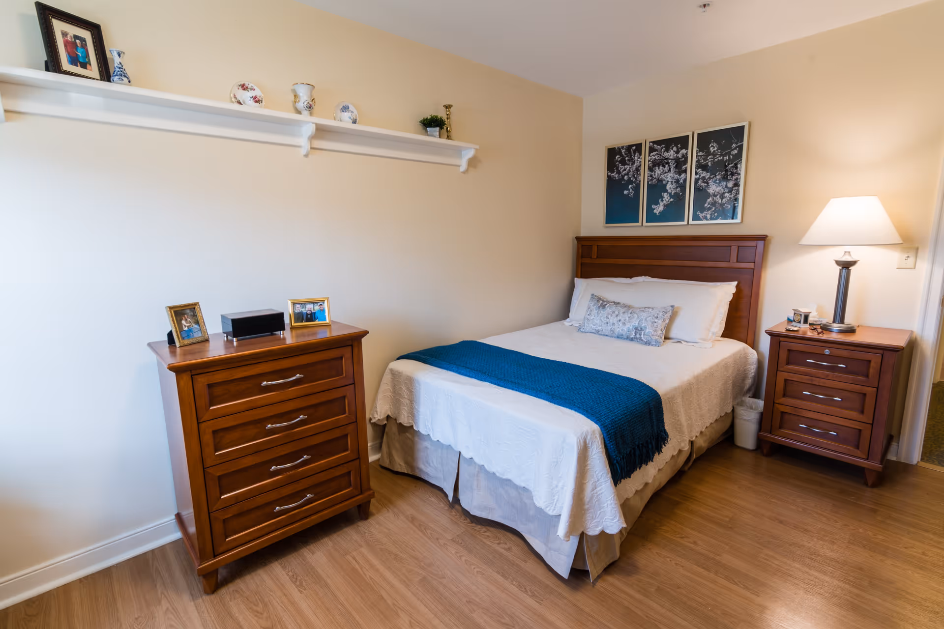 A cozy bedroom with a wooden bed frame, a white bedspread, a blue throw blanket, and a decorative pillow. There is a wooden nightstand with a lamp and a small trash bin next to the bed. On the opposite side, there is a wooden dresser with framed photos and a small black box on top. Above the dresser, a white shelf holds decorative plates and a small plant. The walls are light-colored, and there is a three-panel artwork featuring floral designs above the bed.
