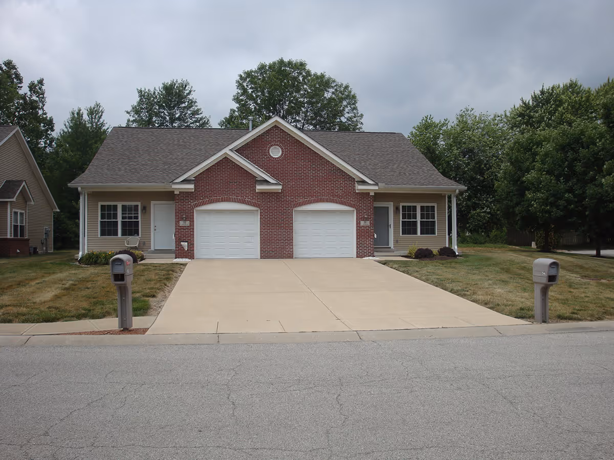 A duplex residential building with two single-car garages, two front doors, and two mailboxes on a paved street. The building has a brick facade in the center and beige siding on the sides, surrounded by grass and trees under a cloudy sky.
