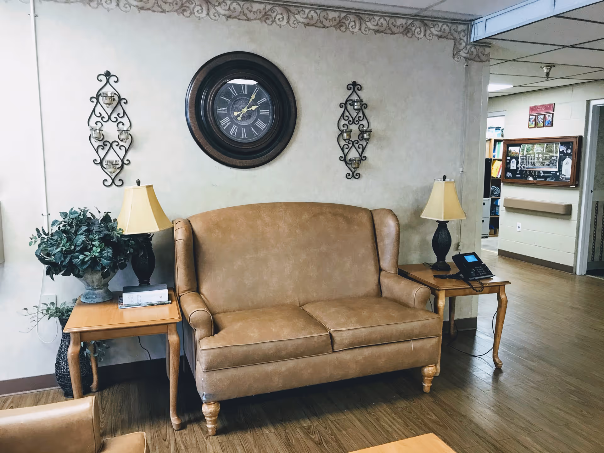 A cozy seating area with a brown leather loveseat flanked by two wooden side tables, each holding a lamp. A large round wall clock hangs above the loveseat, with decorative wrought iron candle holders on either side. A potted plant is placed on the left side table. The room has wood flooring and light-colored walls with a decorative border near the ceiling. In the background, there is a hallway with open doors and a bulletin board on the wall.