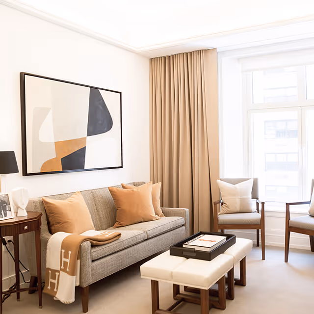 A bright and cozy living room with a gray sofa adorned with beige and brown pillows, a beige throw blanket with the letter H draped over the armrest, two beige cushioned chairs with pillows, a white cushioned ottoman with a black tray on top, a wooden side table with a black lamp and decorative items, beige curtains, and a large abstract painting on the wall.