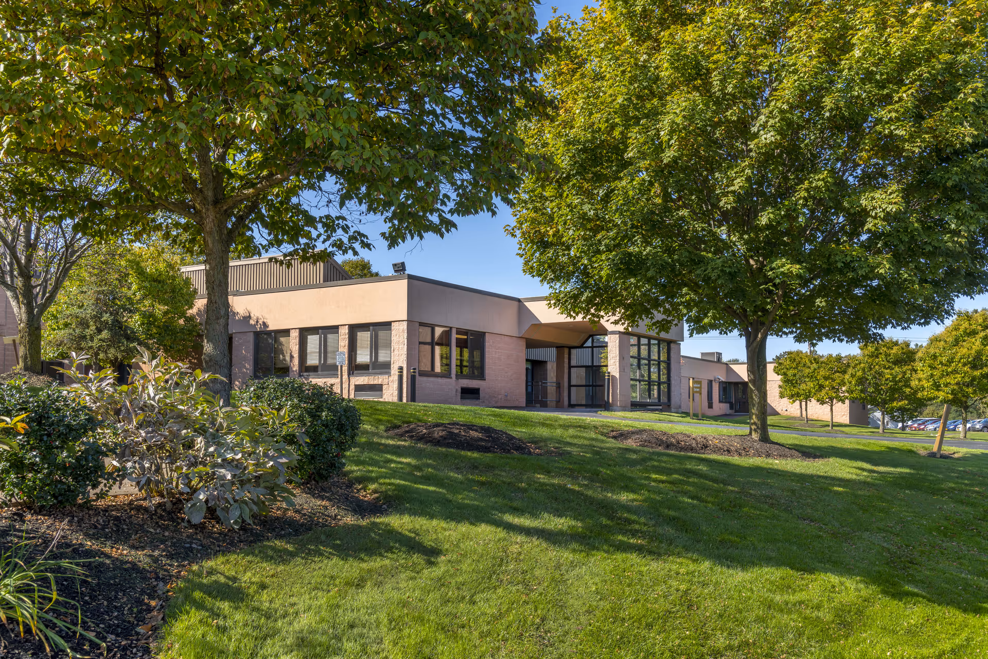 Exterior view of a single-story building with large windows, surrounded by green grass, trees, and shrubs under a clear blue sky.