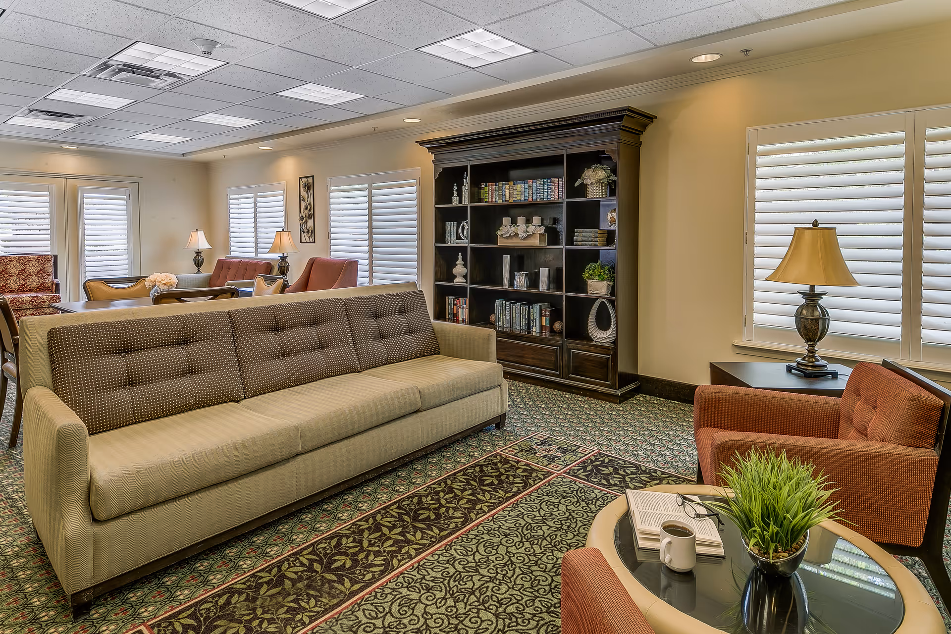A cozy living room area in a senior living facility featuring a long beige sofa with a patterned backrest, two red armchairs, a round glass coffee table with a plant, an open book, and a coffee mug. The room has large windows with white shutters, a dark wooden bookshelf filled with books and decorative items, and several table lamps providing warm lighting. The floor is carpeted with a patterned rug.