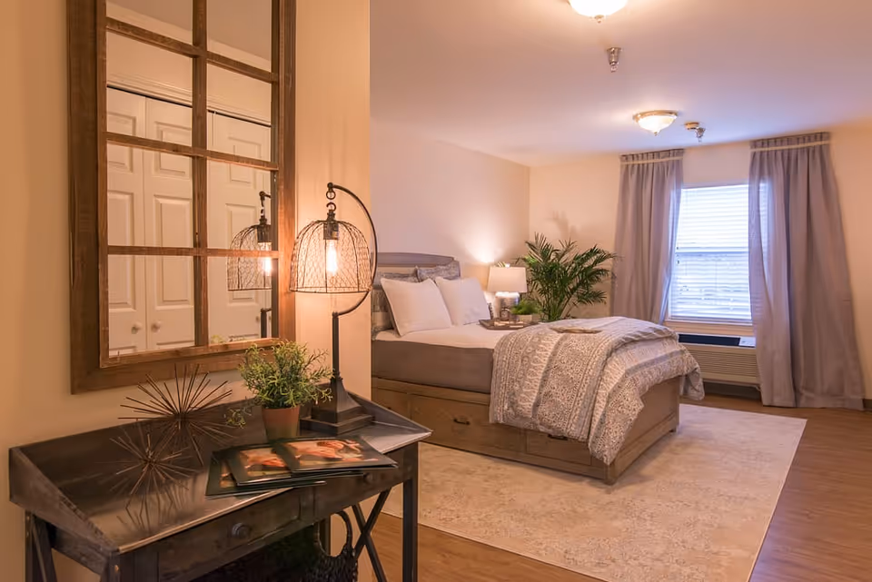 A cozy bedroom with a neatly made bed featuring white pillows and a patterned comforter. There is a wooden desk with a decorative lamp, a small plant, and a photo frame. A large window with curtains allows natural light into the room. The floor is wooden with a light-colored area rug, and a green plant is placed near the bed.